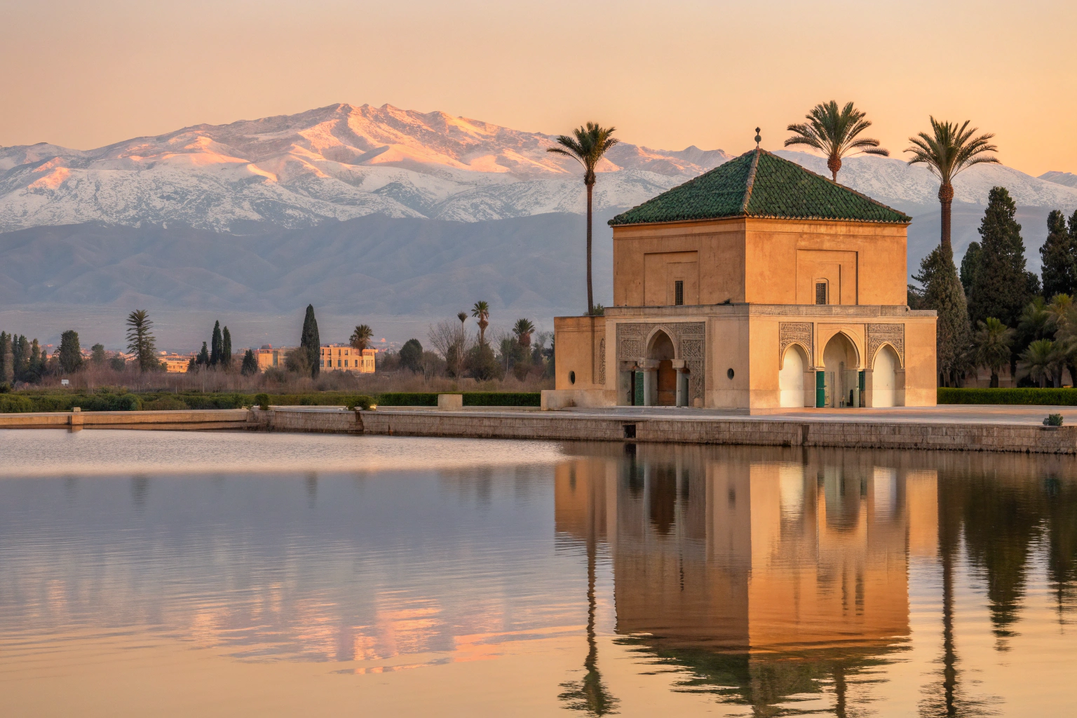 Sunset view of Menara Gardens Marrakech with pavilion reflection in water and Atlas Mountains glowing in the background.