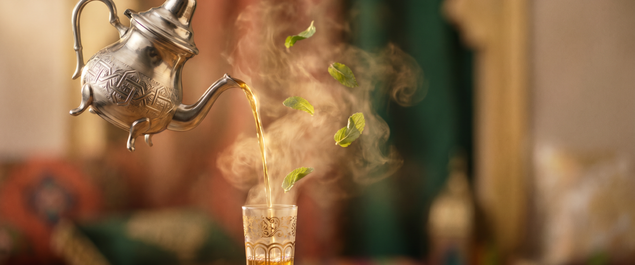 Cinematic image of a silver Moroccan teapot pouring tea in a graceful arc with warm golden light, soft steam, and floating mint leaves against a minimal terracotta and green background.