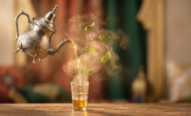 Cinematic image of a silver Moroccan teapot pouring tea in a graceful arc with warm golden light, soft steam, and floating mint leaves against a minimal terracotta and green background.