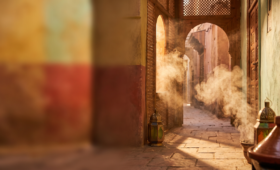 Golden sunset light streaming through a traditional Marrakech archway with warm spice-toned hues and soft drifting steam, evoking the sensory atmosphere of a Moroccan food tour.