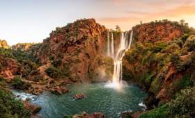 Panoramic view of Ouzoud Waterfalls in Morocco with multi-tiered cascades, red rock cliffs, lush olive trees, and a small raft near the base during golden hour.