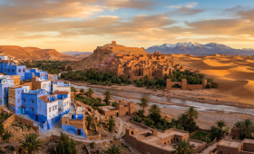 Panoramic banner image of a traditional Moroccan village featuring blue-painted houses, an ancient earthen kasbah, and desert dunes in the distance, surrounded by Atlas Mountains under a warm golden sunset sky.