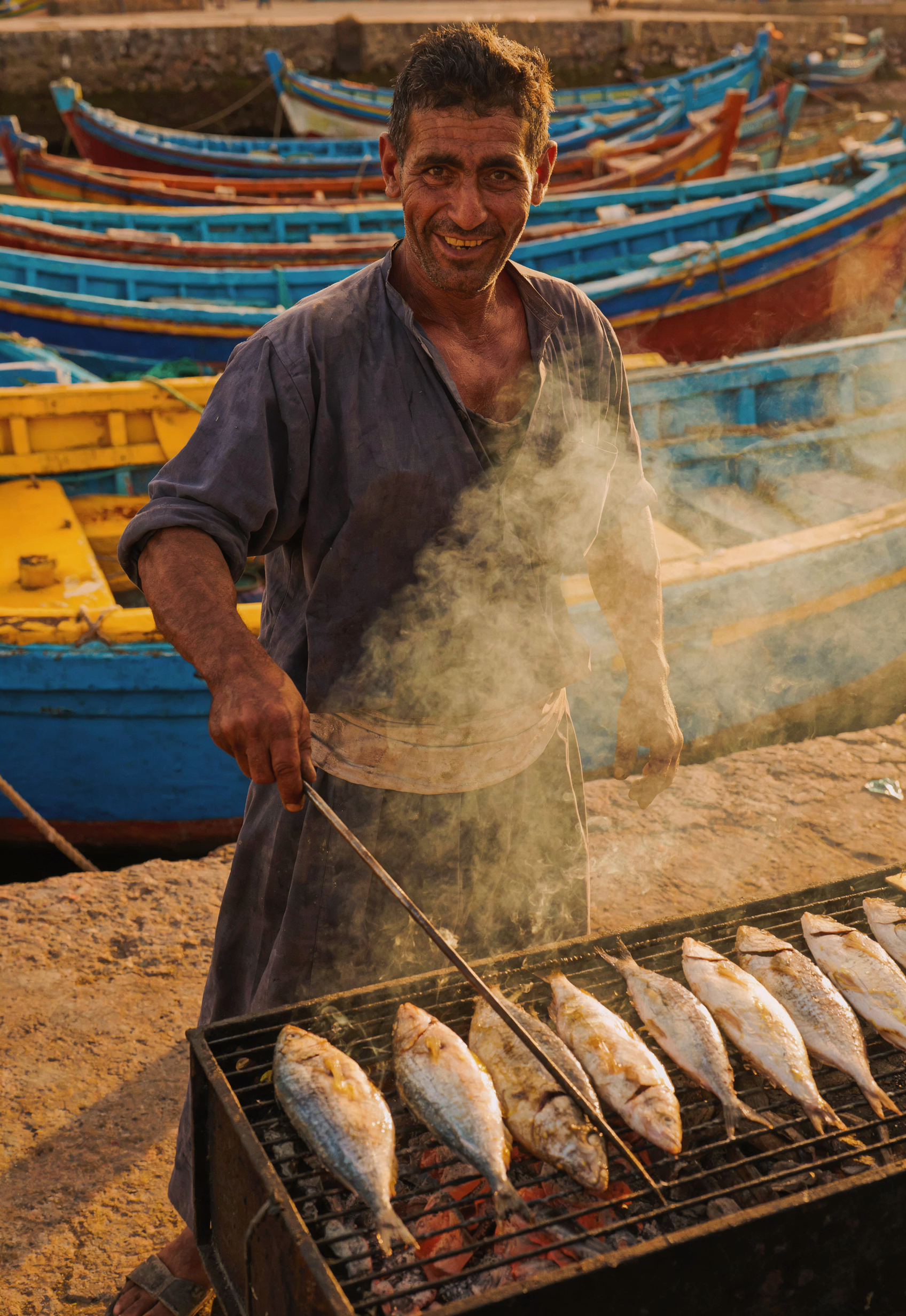 Fresh grilled fish at Taghazout Morocco beach fisherman port, local man cooking, colorful boats, authentic Moroccan coastal food culture