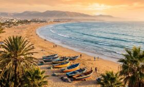 Golden hour panoramic view of Agadir beach with palm trees, fishing boats, and Atlas mountains - affordable Morocco beach holidays