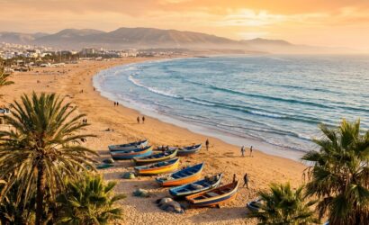 Golden hour panoramic view of Agadir beach with palm trees, fishing boats, and Atlas mountains - affordable Morocco beach holidays