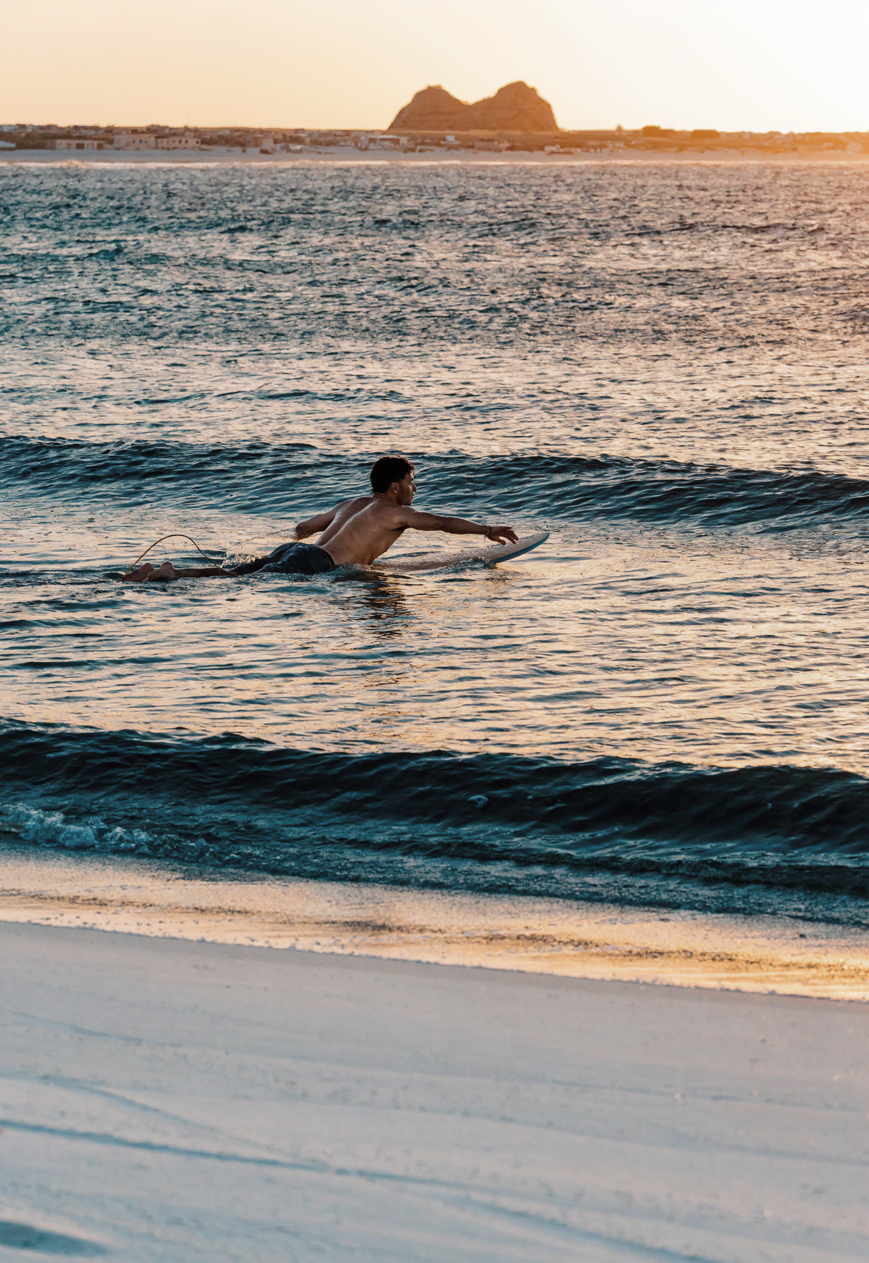Surfer at sunrise on Taghazout Morocco white beach, golden light on Atlantic waves, Camel Rock silhouette, peaceful surf culture scene