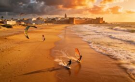 Essaouira Morocco beach panoramic sunset view with surfers and kitesurfers, golden sand and Atlantic waves, historic medina skyline, perfect Morocco beach vacation destination
