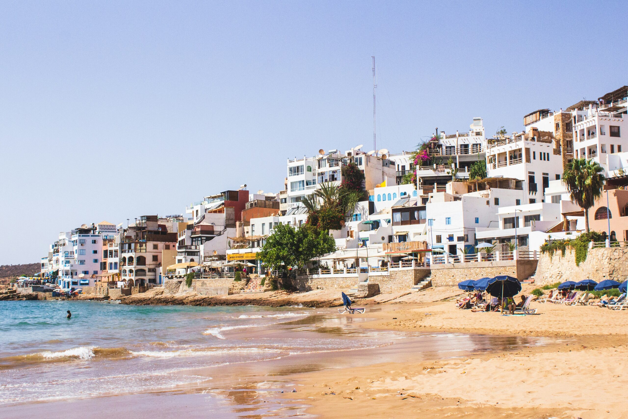 A beach scene with a large body of water, a sandy shore, and a city in the background, Agadir