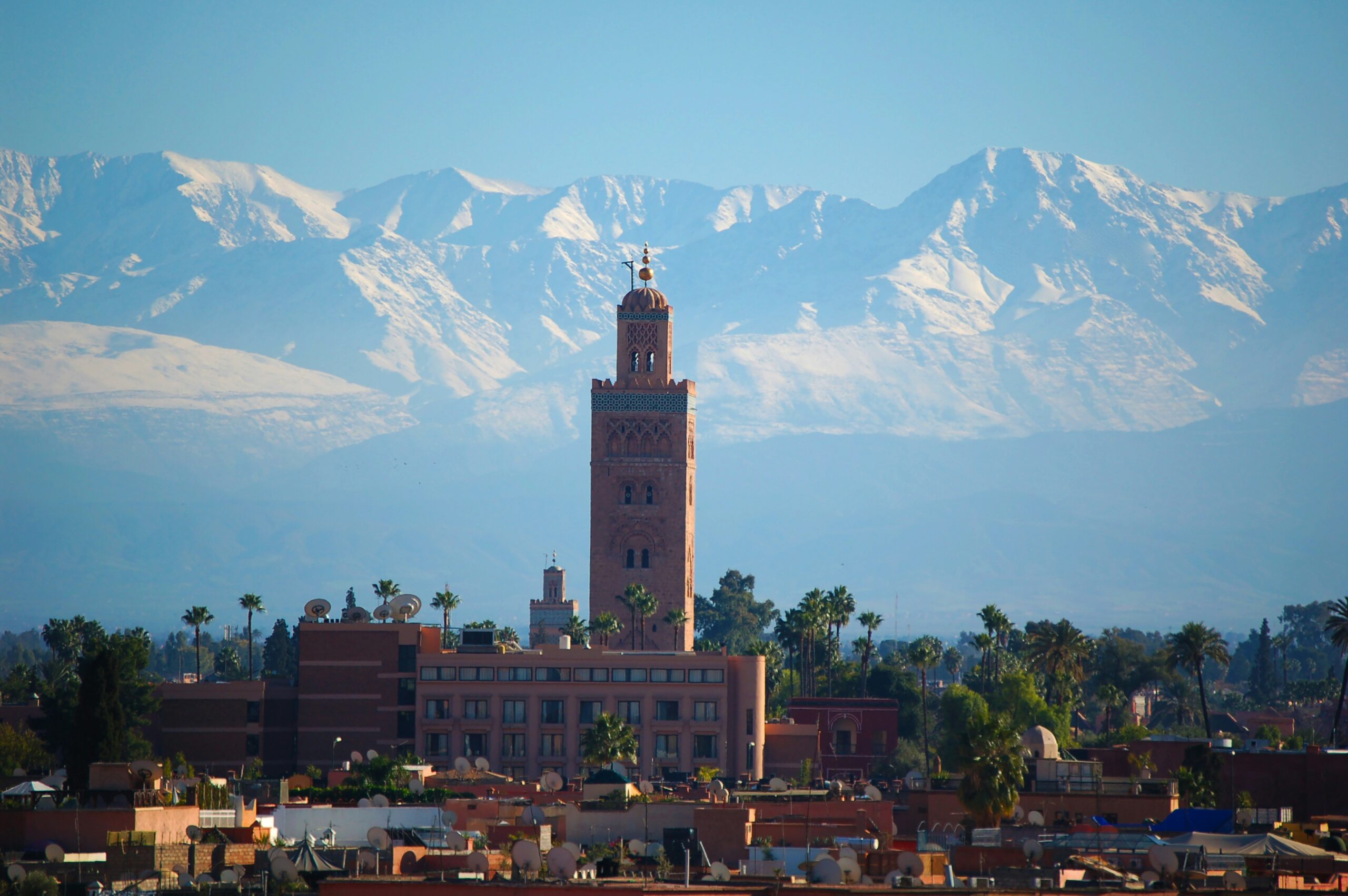 A tall, ornate building with a pointed roof, situated in a city with snow-capped mountains in the background, Marrakech